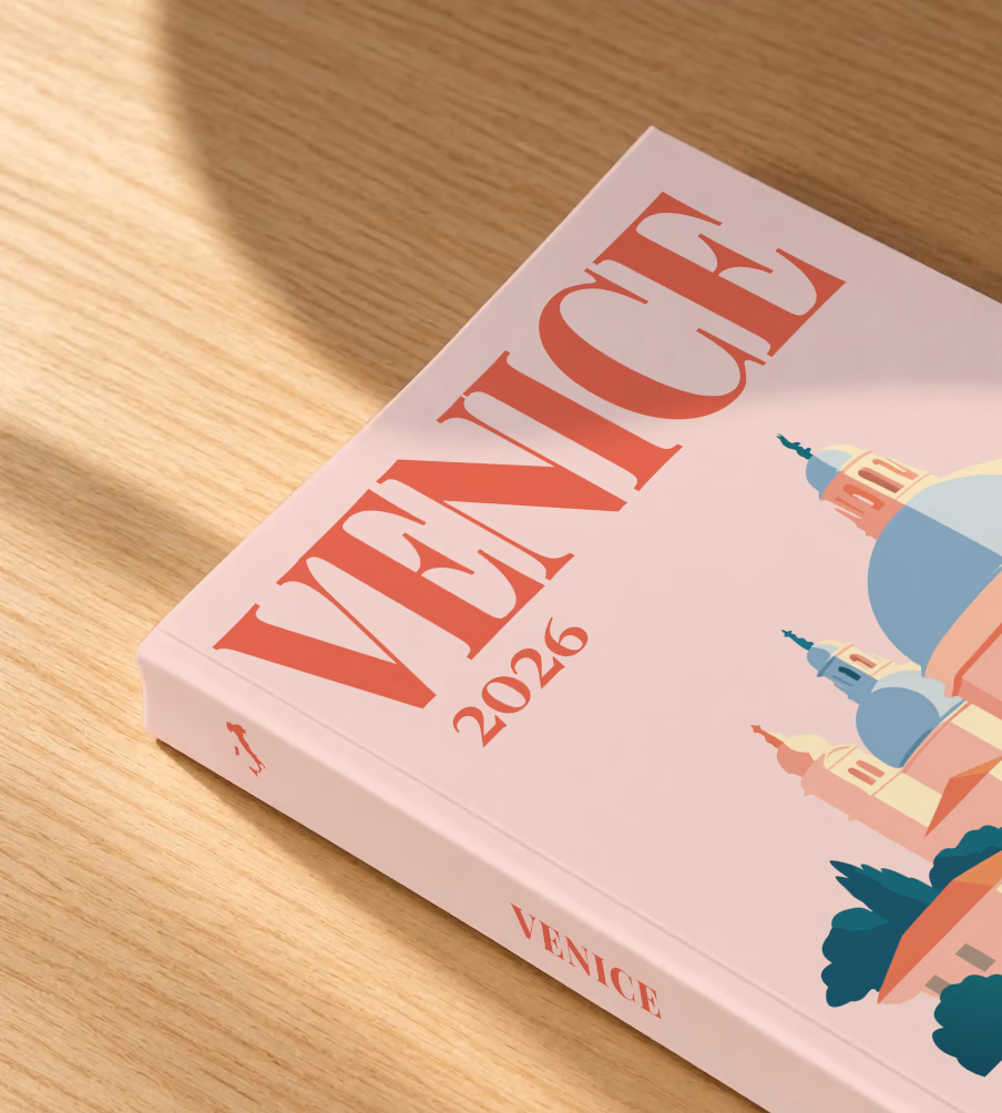 Venice softcover photo book with the illustrated cover of Santa Maria della Salute on blush pink, lying on an oak desk in dappled afternoon sunlight