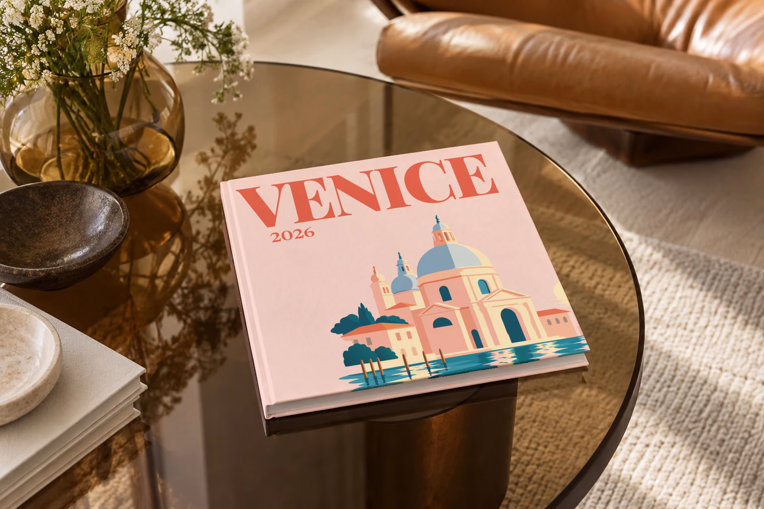 Venice square hardcover photo book with the illustrated cover of Santa Maria della Salute on blush pink, on a smoked glass coffee table beside a leather armchair in warm afternoon light