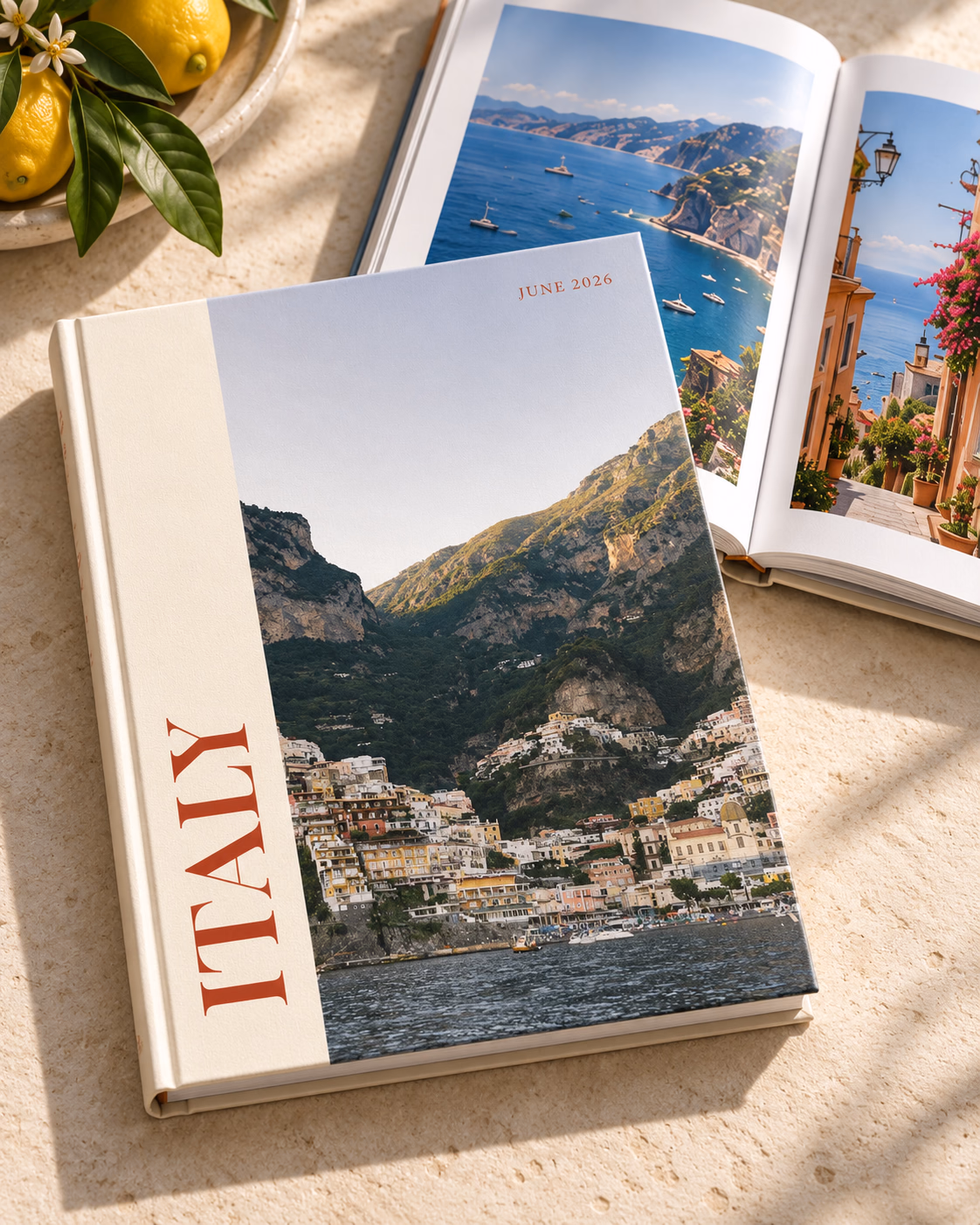 Italy photo book with the Editorial cover, a photo of a cliffside coastal town beside ITALY in red on a cream panel, on a stone table next to an open book of coastal photos and a bowl of lemons