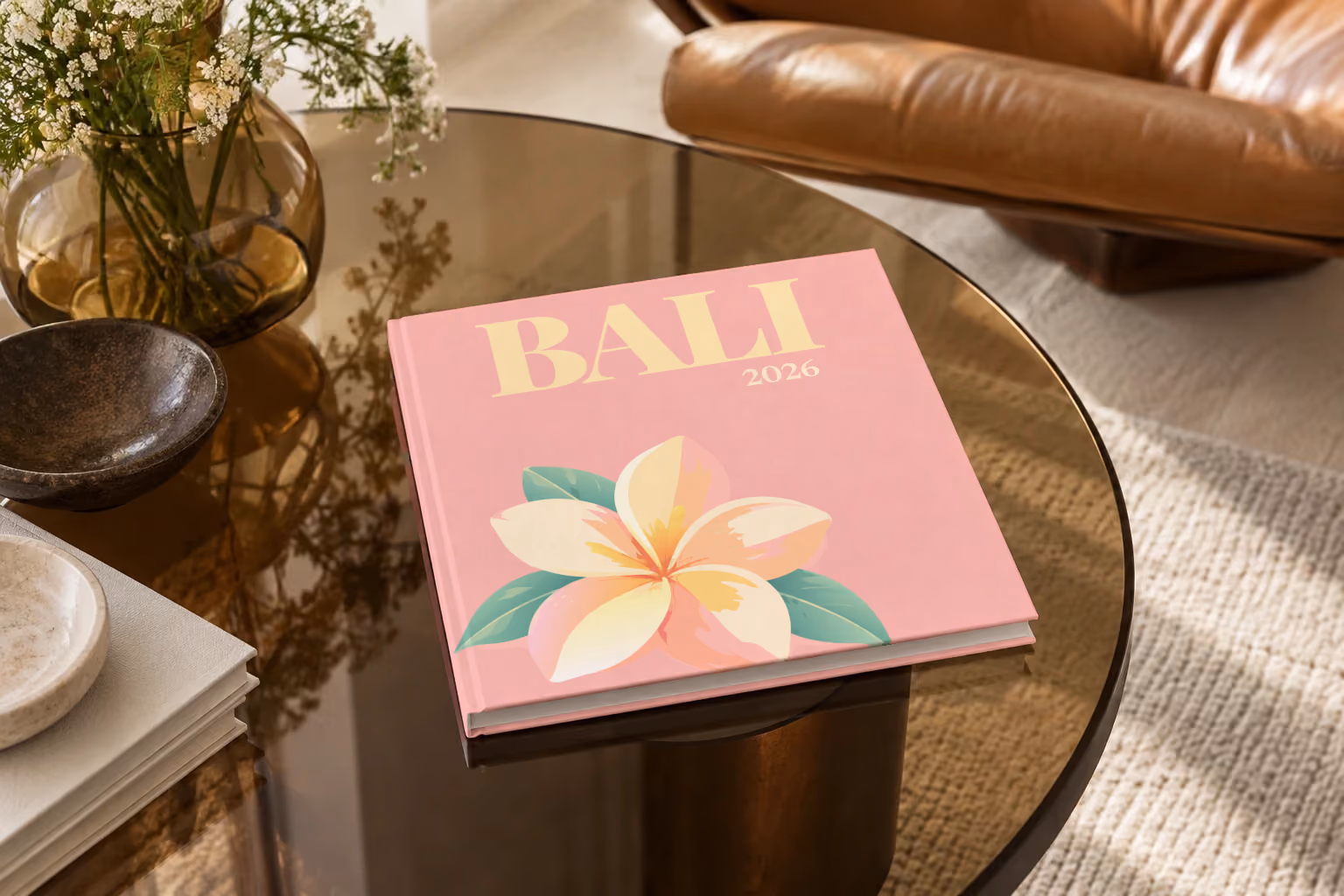 Bali hardcover photo book with an illustrated cover of a frangipani flower, on a smoked glass coffee table beside a leather armchair in warm afternoon light