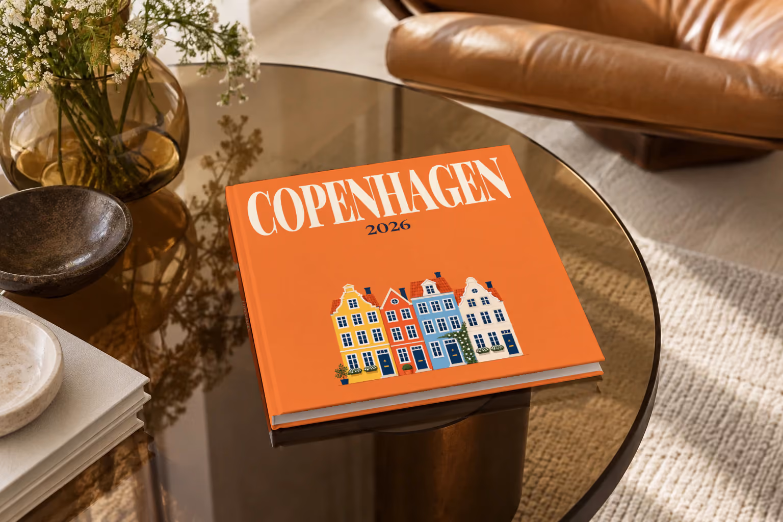 Copenhagen hardcover photo book with an illustrated cover of the colourful Nyhavn houses, on a smoked glass coffee table beside a leather armchair in warm afternoon light