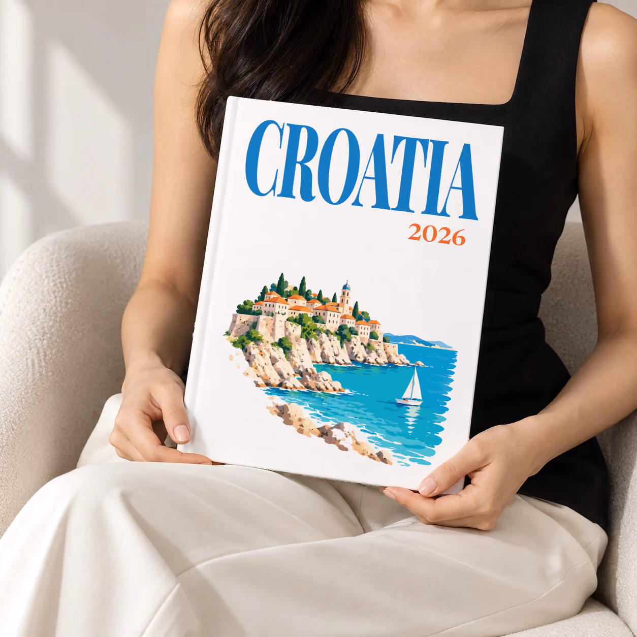 Croatia hardcover photo book with an illustrated cover of an Adriatic old town and sailing boat, held upright in two hands by a woman in a black top, sitting in a cream boucle armchair