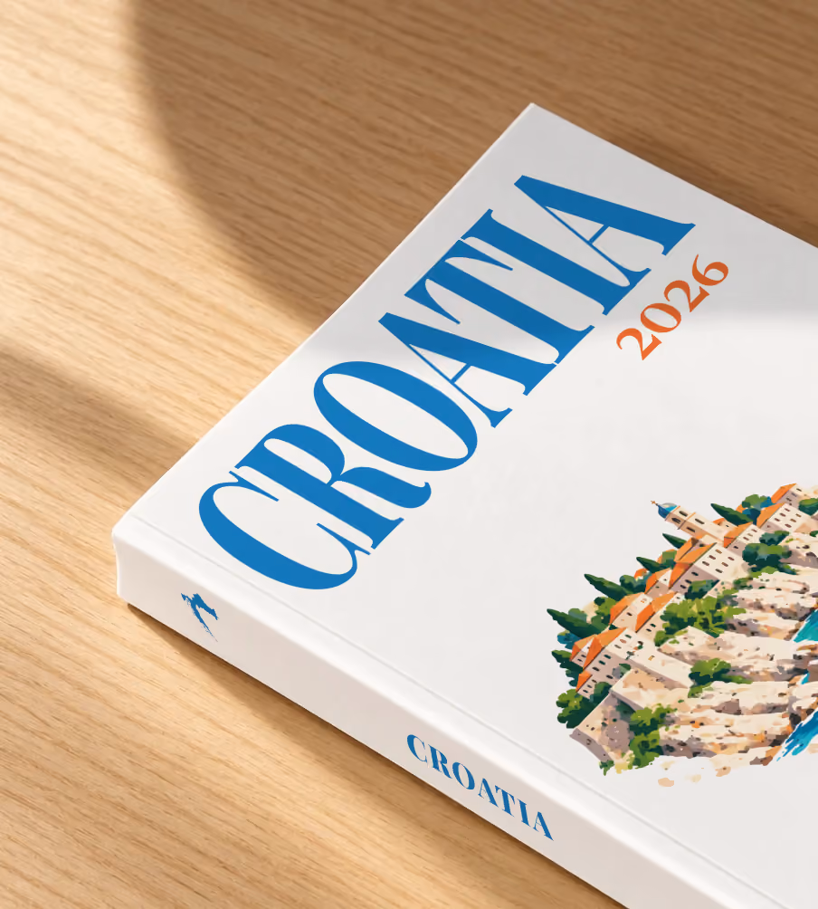 Croatia softcover photo book with an illustrated cover of an Adriatic old town and sailing boat, lying on an oak desk in dappled afternoon sunlight
