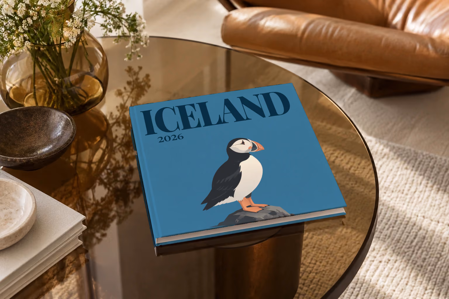 Iceland hardcover photo book with an illustrated cover of a puffin on a rock, on a smoked glass coffee table beside a leather armchair in warm afternoon light