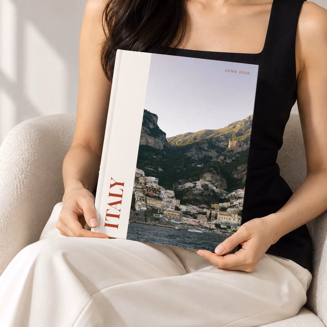 Italy hardcover photo book with the Editorial cover, held upright in two hands by a woman in a black top, sitting in a cream boucle armchair