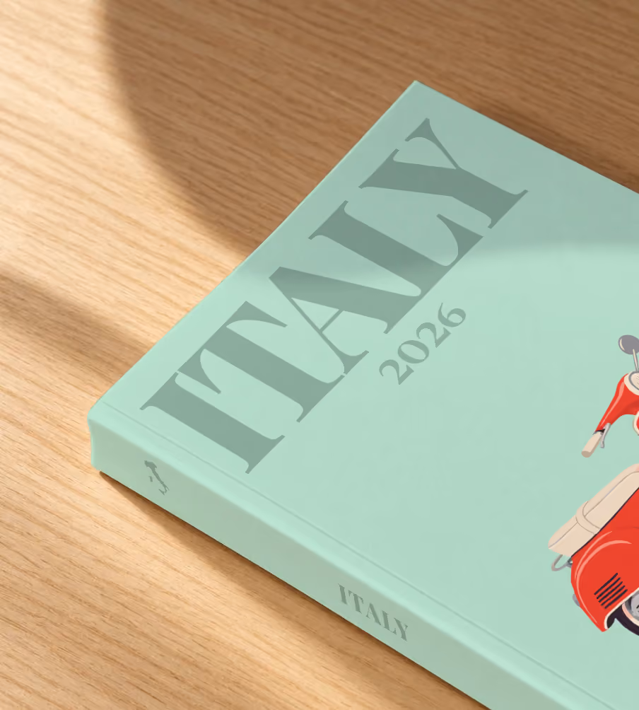 Italy softcover photo book with an illustrated cover of a red Vespa scooter on mint green, lying on an oak desk in dappled afternoon sunlight