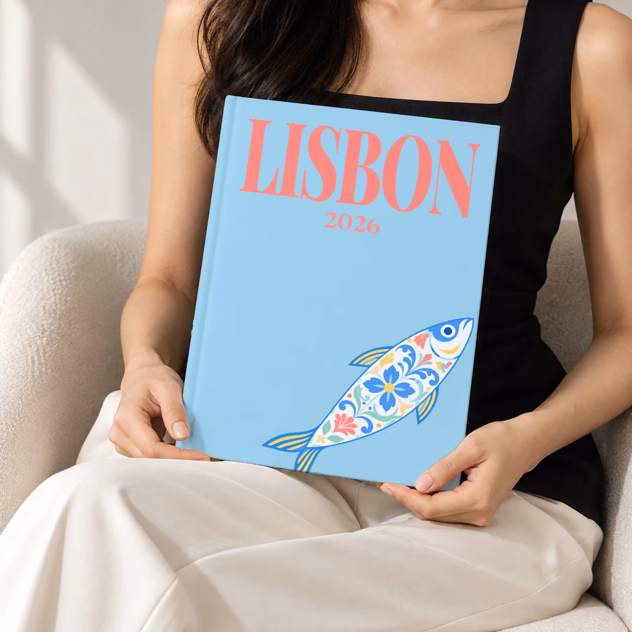 Lisbon hardcover photo book with an illustrated cover of a sardine painted like azulejo tiles, held upright in two hands by a woman in a black top, sitting in a cream boucle armchair
