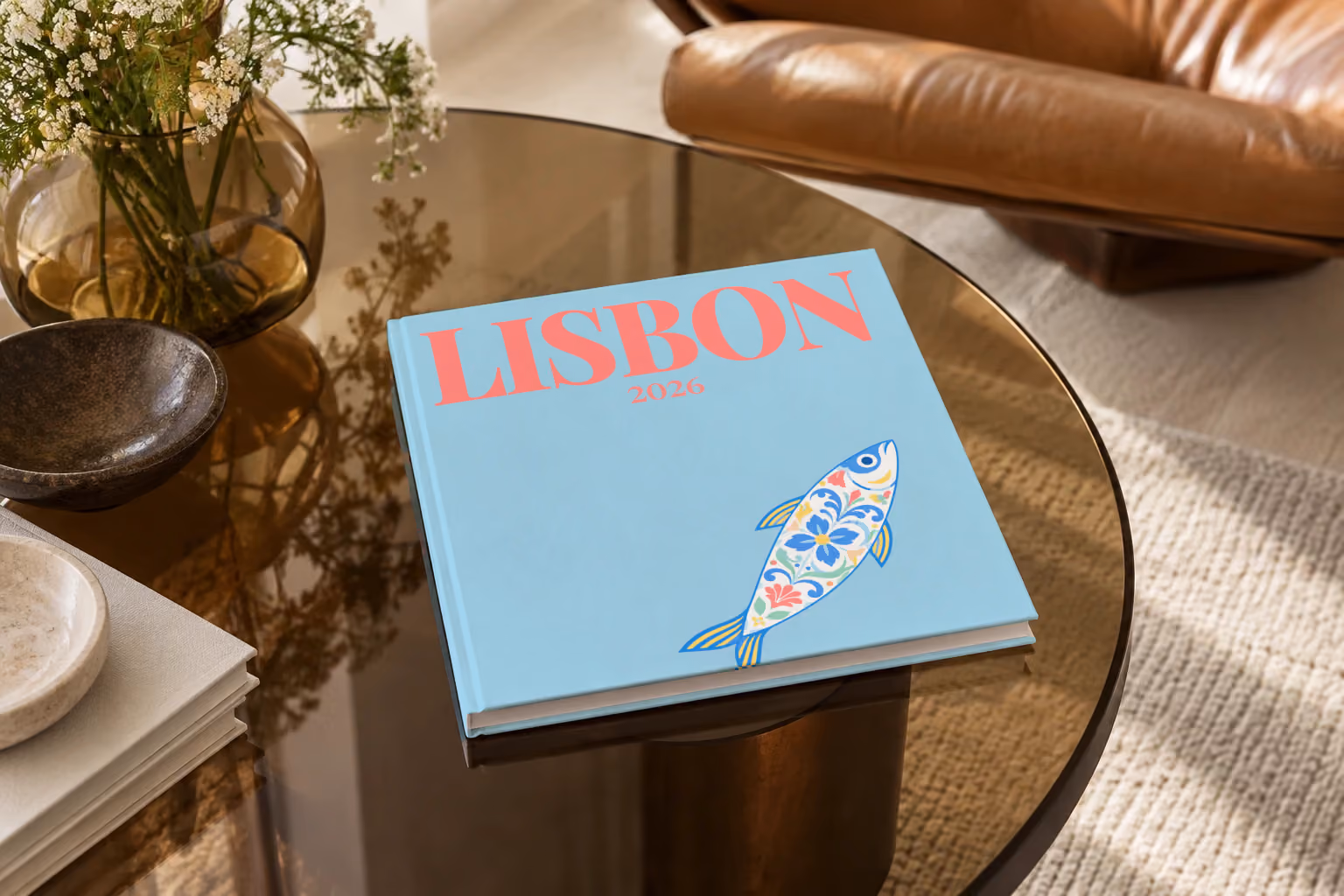 Lisbon hardcover photo book with an illustrated cover of a sardine painted like azulejo tiles, on a smoked glass coffee table beside a leather armchair in warm afternoon light