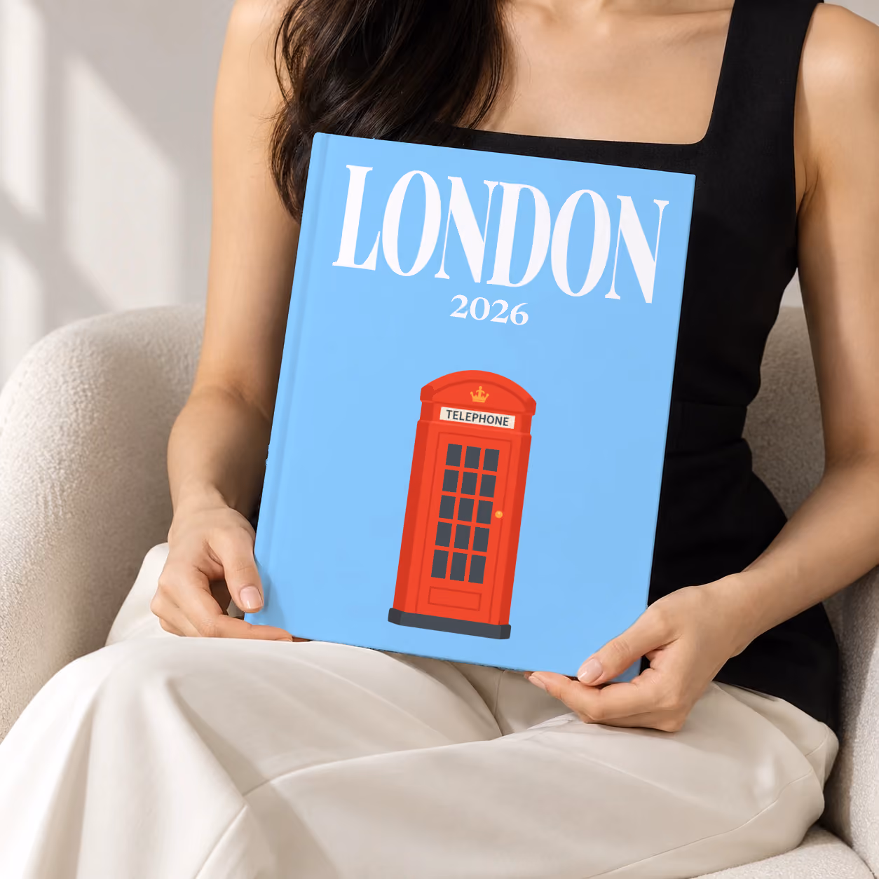 London hardcover photo book with an illustrated cover of a red telephone box, held upright in two hands by a woman in a black top, sitting in a cream boucle armchair