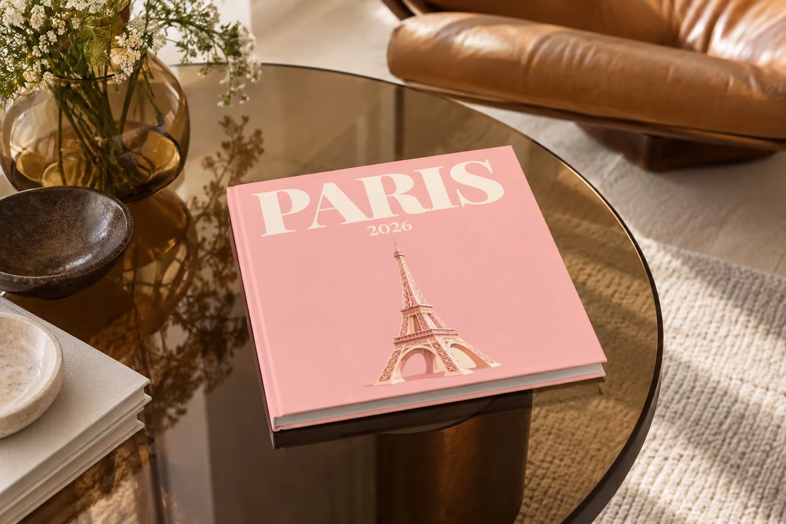 Paris hardcover photo book with an illustrated cover of the Eiffel Tower on blush pink, on a smoked glass coffee table beside a leather armchair in warm afternoon light