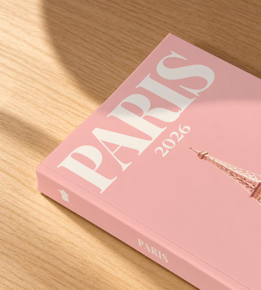 Paris softcover photo book with an illustrated cover of the Eiffel Tower on blush pink, lying on an oak desk in dappled afternoon sunlight