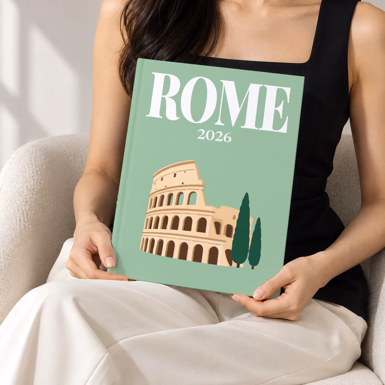 Rome hardcover photo book with an illustrated cover of the Colosseum and cypress trees, held upright in two hands by a woman in a black top, sitting in a cream boucle armchair
