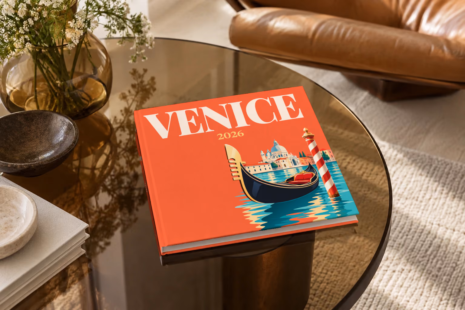 Venice hardcover photo book with an illustrated cover of a gondola, a striped mooring pole and Santa Maria della Salute, on a smoked glass coffee table beside a leather armchair in warm afternoon light