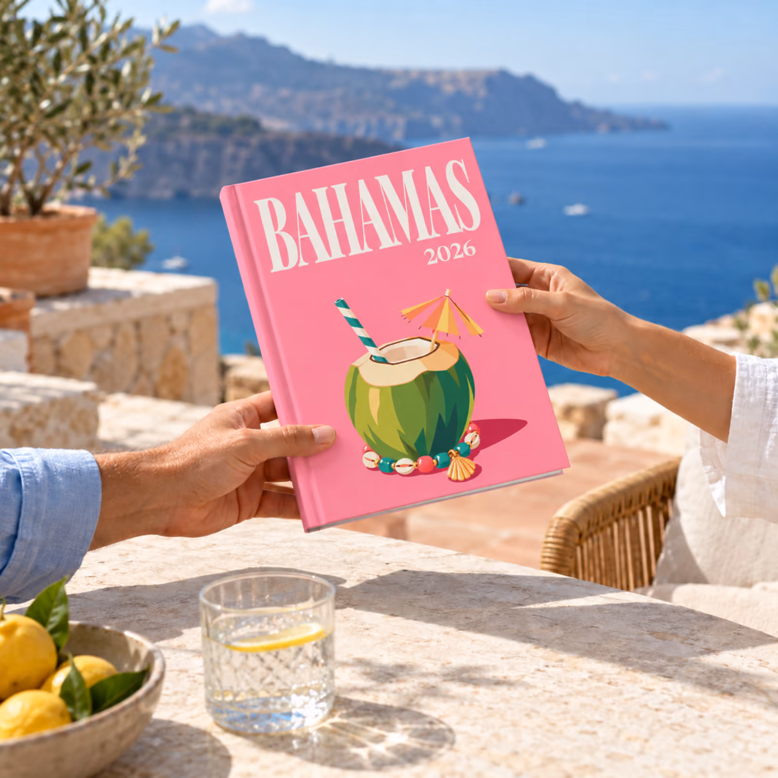 Two people passing a Bahamas travel photo book across a terrace table above the sea