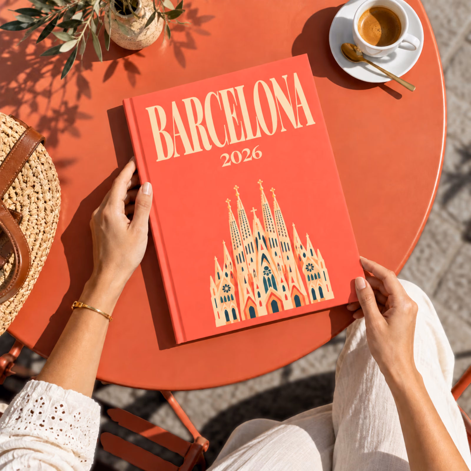 Hands holding a red Barcelona travel photo book illustrated with the Sagrada Família, on a café table with a coffee