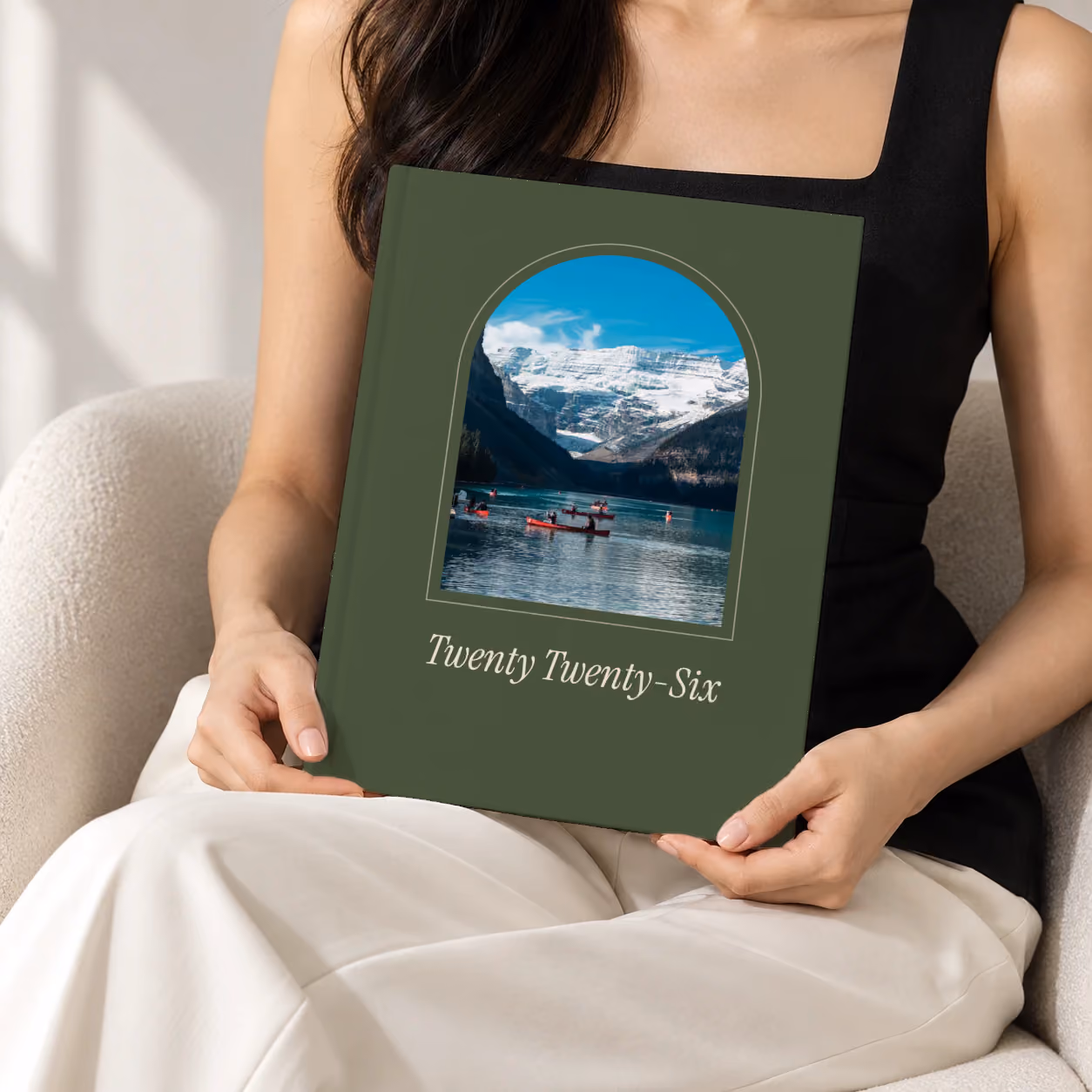 Arch year in review hardcover photo book, an olive cover with a photo framed in an arch, held upright in two hands by a woman in a black top, sitting in a cream boucle armchair