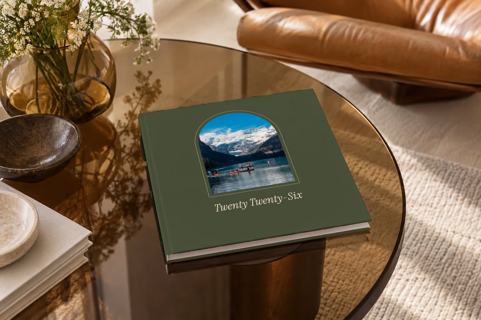 Arch year in review square hardcover photo book, an olive cover with a photo framed in an arch, on a smoked glass coffee table beside a leather armchair in warm afternoon light