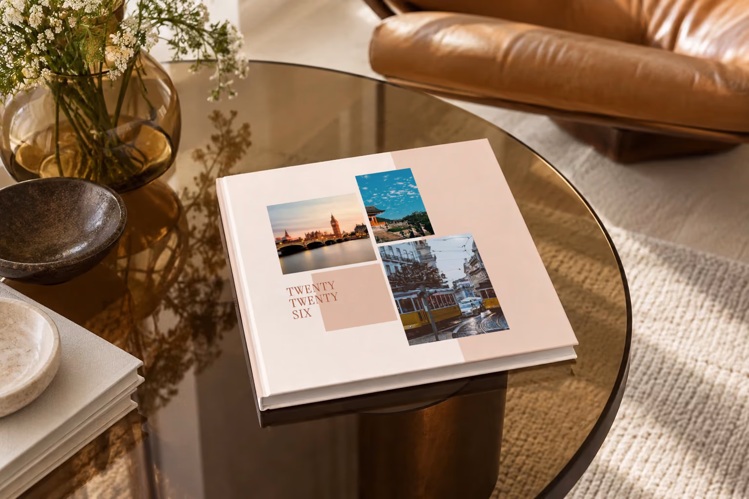 Editorial II year in review square hardcover photo book, a blush editorial cover with three travel photos, on a smoked glass coffee table beside a leather armchair in warm afternoon light