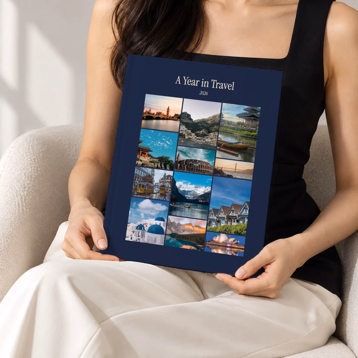 Mosaic III year in review hardcover photo book, a navy cover with a mosaic of travel photos, held upright in two hands by a woman in a black top, sitting in a cream boucle armchair