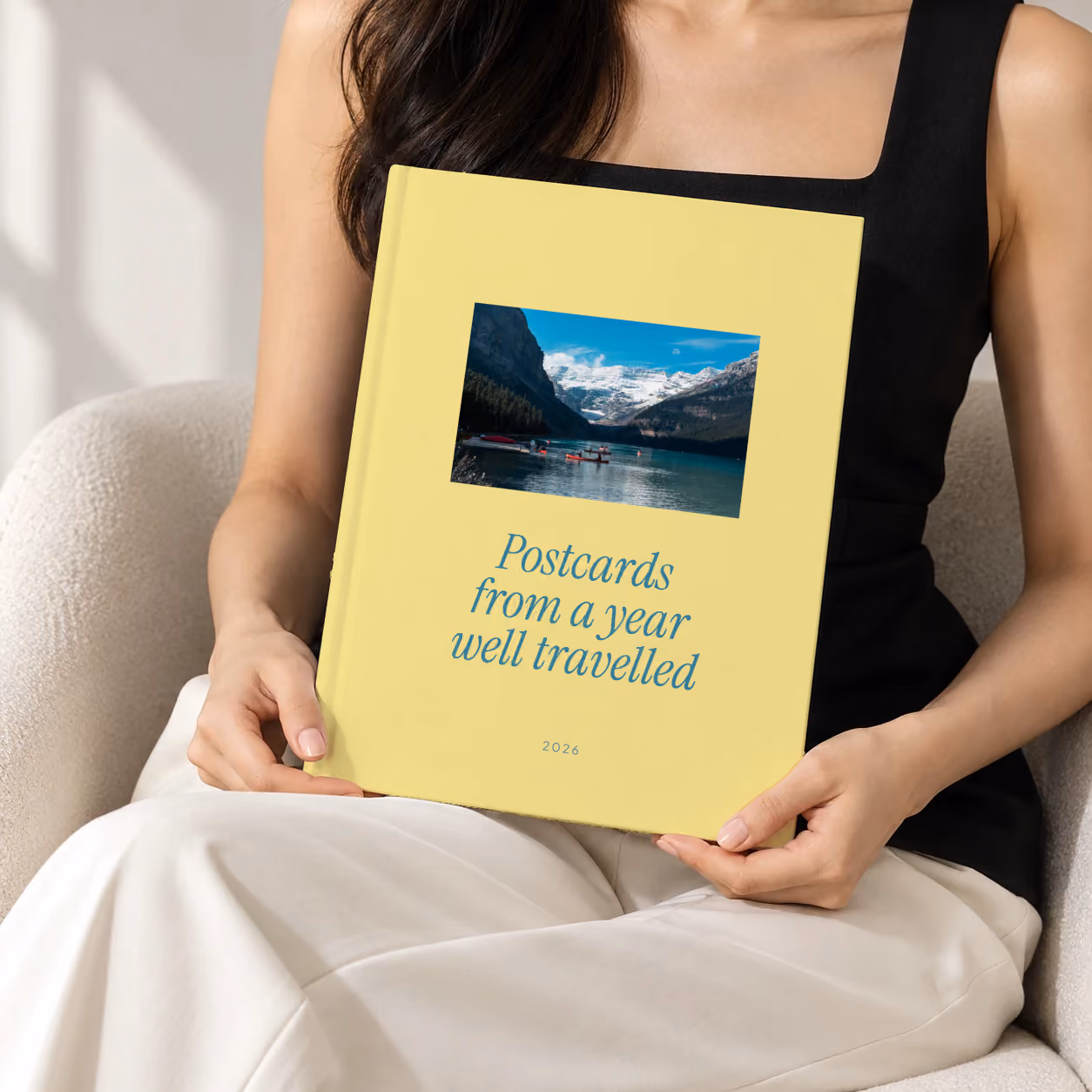 Postcards year in review hardcover photo book, a yellow cover reading Postcards from a year well travelled, held upright in two hands by a woman in a black top, sitting in a cream boucle armchair