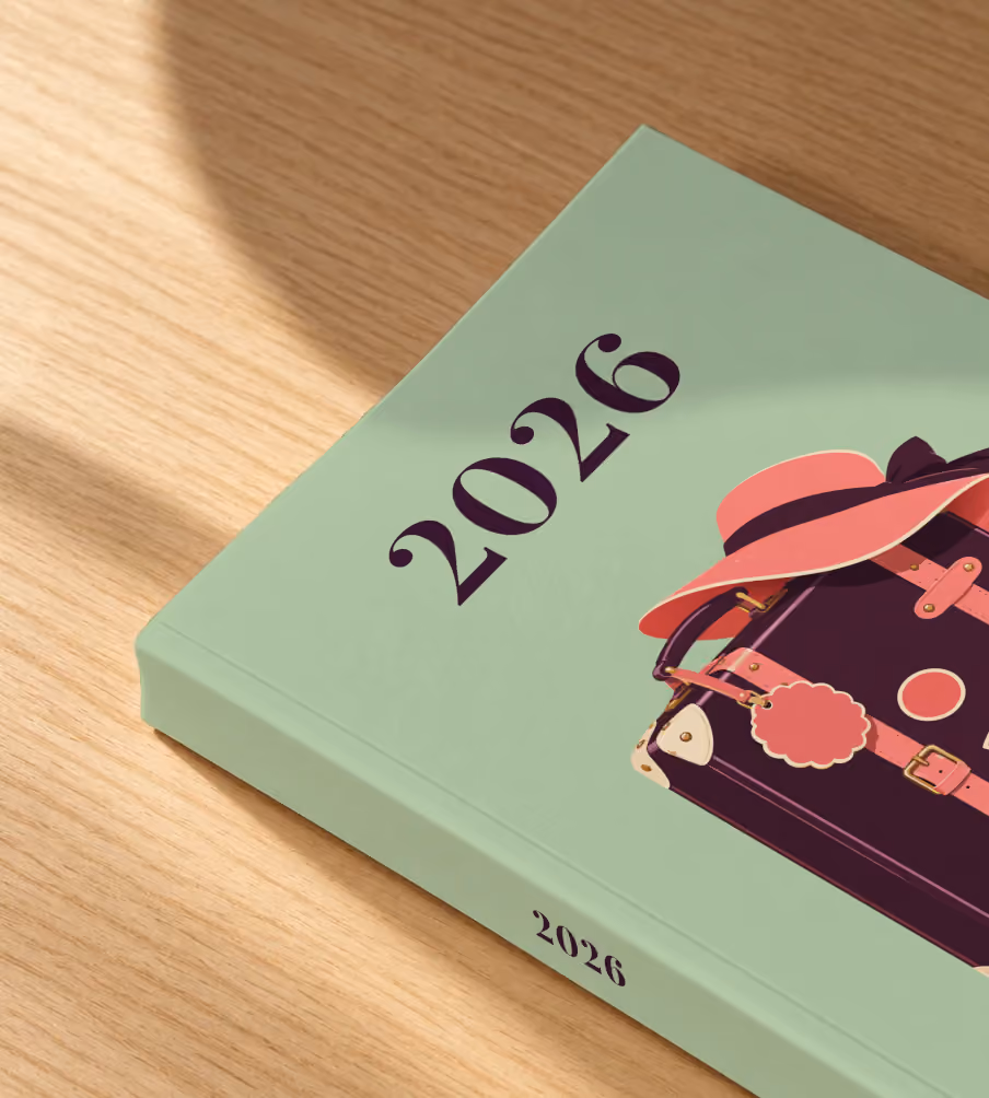 Suitcase year in review softcover photo book, a sage cover with an illustrated suitcase and sun hat, lying on an oak desk in dappled afternoon sunlight