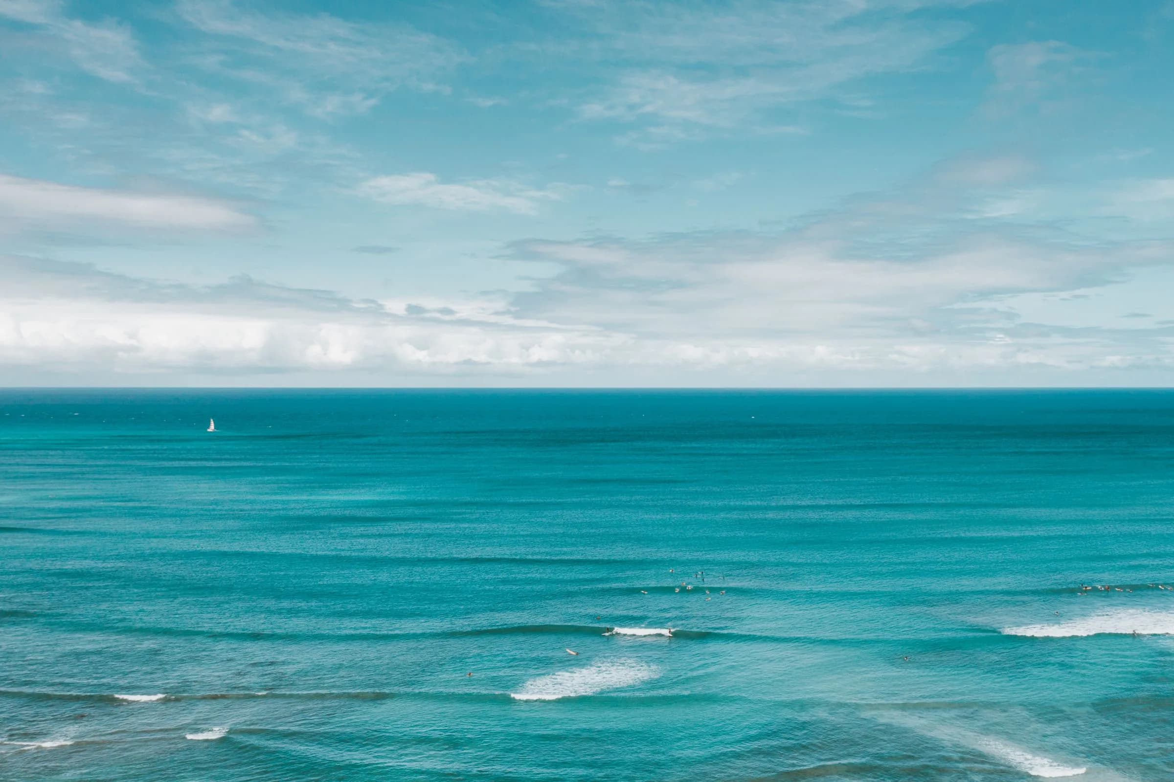 Turquoise ocean beneath a soft blue cloud horizon with a distant sailboat