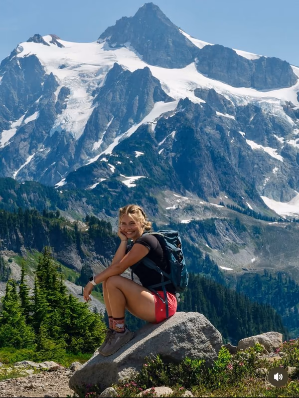 Backpacker resting beside a mountain trail with snowy peaks behind her